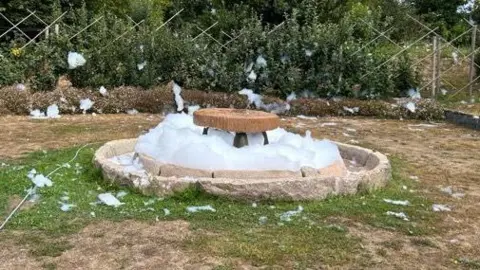 Christopher Beaumont A fountain filled with foam with grass around it and a fence behind.