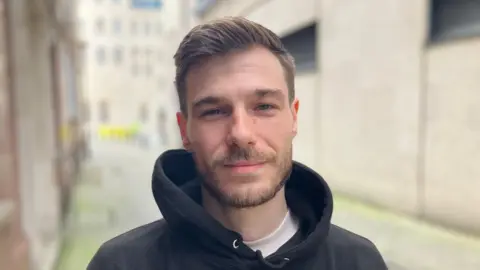 BBC Close-up of Tyler with alley and house in background. He has a brown beard and short brown hair and is wearing a black hoodie with a white t-shirt underneath. 