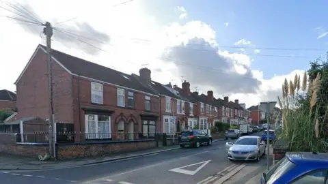 A set of red-brick terraced homes with cars parked alongside the curb on either side of the road.