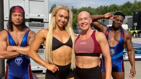 Poppy Bulmer A female 'contestant' smiles joyfully, alongside three gladiators at an outdoor/ backstage location. She has a bald head, an athletic body and wears a crop top and shorts. The first male gladiator has long black hair and facial hair wears a shiny blue and red body suit and a red bandana. His arms are crossed and he has a menacing expression. The second male gladiator has short black hair and a muscular frame, and wears the same suit. He looks friendly and makes the 'V' peace sign with his hand.  The third gladiator is a tall female with long blond hair and a big smile. She has an athletic body and wears black shorts and a black bikini style top.