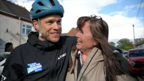 A man with a blue cycle helmet and black top hugging a woman in a grey coat with long brown hair