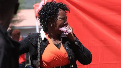 A distressed woman in an orange top and black jacket looks to the side holding a handkerchief to her face.