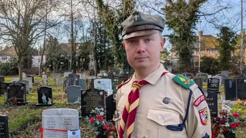 Lukasz Lakomy wearing the Polish Scouting Association uniform and standing in a cemetery. He is wearing a peaked cap in khaki and a light brown shirt with badges. A yellow and red tie is around his neck. Behind him are black, white and grey graves, grass, trees, hedges and beyond that houses.