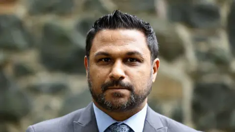 BBC A man in a grey suit, blue shirt and blue tie is looking at the camera. He has dark hair and a short dark beard
