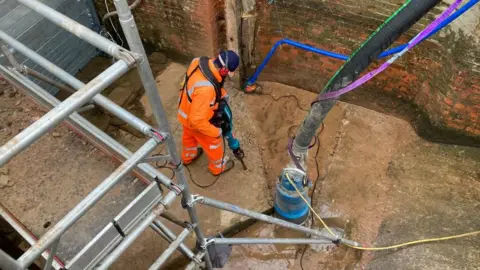 Alex Pope/BBC A man, in full orange high-viz clothing, holding a drill, down the bottom of a lock, with machinery, and metal scaffolding by him. There are red brick walls around him. 
