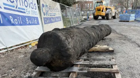 The photo shows a black iron cannon that is laid on a wooden pallet on the ground. Behind it is an orange excavator. There is metal fencing to the left, with white and blue banners reading 'CR Reynolds' on them. 