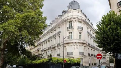 AFP via Getty Images A large, traditional Parisian building on the corner of a street, built from pale stone. It has about six floors, some with balconies. Green trees line the street. 