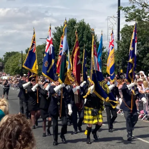 Several people dressed in dark blue military outfits holding up flags with the union jack on and parading down a street. There are people on railings either side watching them.