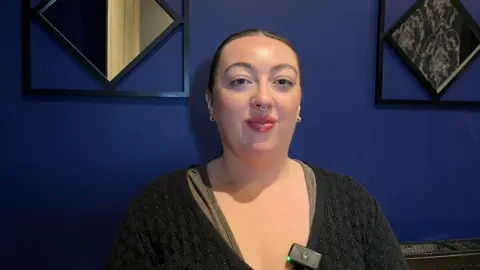 Abigail Grearson is sitting against a blue background. She has dark hair swept back. 