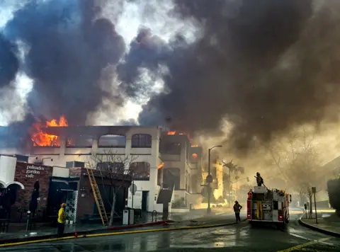 Jeff Gritchen / MediaNews Group / Orange County Register Firefighters battle a fire at an apartment complex on La Cruz Drive during the Palisades Fire on Wednesday, with flames and smoke billowing from the structure.