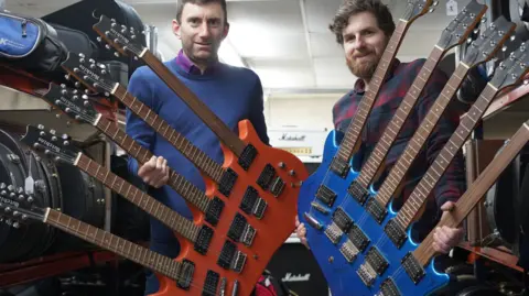 Gardiner Houlgate Two five neck guitars, one orange the other blue being held by two men looking at the camera with a Marshall amplifier in the background