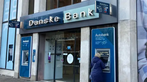 A picture of the front of a Danske Bank branch in Belfast city centre. Cash machines are on either side of the automatic glass doors and the sign is in both blue and white.