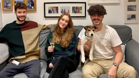 Caleb Tutt Three people sat on a grey sofa with framed picture behind them. On the left a man smiles with a dark brown goatee and short brown hair. His arms are outspread and he has a green, light brown and terracotta jumper on with navy joggers, his phone is on his lap. In the middle a woman smiles with long, light brown hair. She holds a flute of champagne and has a striped green and blue jumper on with dark trousers. On the right is a man with curly brown hair, also smiling. He holds a dog in his right hand, and has a white t-shirt and beige trousers on. 