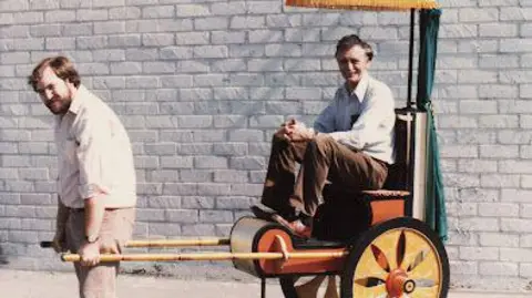 Supplied Two men are posing with a custom-built, two-wheeled vehicle. One man sits in the vehicle while the other stands in front of it, holding the poles. 