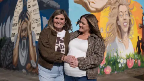 Wendy Robinson pictured with her daughter Amy standing in front of a colourful mural on two sides of a wall. Amy, on the right, has blonde hair and is smiling. There are blue skies, sun, flowers and birds behind her. On the mural towards the left is a picture of a man holding his head in his hands with words including 'cancer, stress, no, health' written in a lung-diagram on a dark blue background. 
