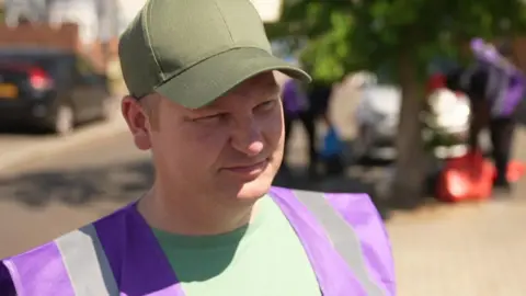 A man in a green cap and purple hi-vis vest stands on a sunlit residential street during a clean-up event.