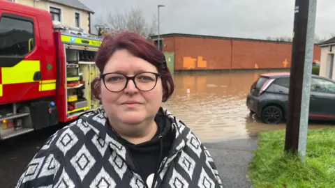 Sarah Kirkwood standing close to flooded homes in Park Avenue, Dundonald on Saturday 23 November.  She has short, red hair and black wide-rimmed glasses.  She is wearing a black hoodie under a black and white diamond-patterned coat. 