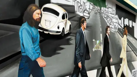 A mural depicting the four band members of The Beatles walking over a zebra crossing. One is in a white suit, two are in dark suits and the fourth is a denim top and jeans. Cars are depicted in the background. 