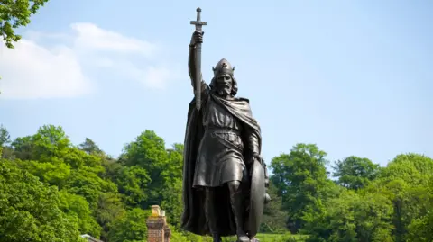 A bronze seen in front of a blue sky and green trees depicts King Alfred the Great holding a sword aloft with his right arm.