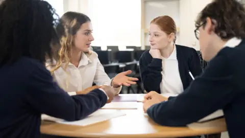 Getty Images A stock image group of young people in a classroom sit around a table during a discussion. The two who have their faces to the camera are female and wear a navy and white school uniform. 