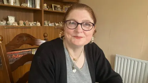BBC Paula is looking at the camera and smiling. Her hair, which is auburn coloured, is pulled back into a pony tail behind her head. She has blue eyes. She has silver hooped earrings in. She is sat on a brown wooden chair. In the background is a brown wooden cabinet with cubby holes. There is lots of different little decor houses on the shelves.