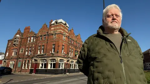 A man with white hair and a beard, wearing a khaki green puffer jacket and a green t-shirt, stands to the right of a red-brick period building.