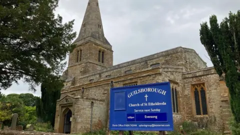 The outside of a church with a blue sign that reads "Guilsborough" at the front