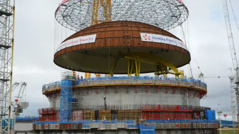 A large copper-coloured dome being lowered into place on top of a big concrete cylinder using a yellow crane. There are steel structures and scaffolding surrounding the cylinder. 
