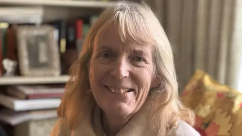 BBC/Genevieve Tudor A woman with shoulder-length fair hair is wearing a white sweater and sitting on a sofa in a living room with a bookshelf and framed photo behind her.