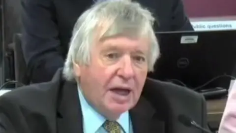 A man with grey hair wearing a suit jacket, shirt and tie pictured from the shoulders up speaking into a microphone. The shoulders of other people sat around him can be seen, including someone on a laptop behind him. 