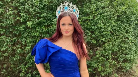 Ryan Cowburn A teenage girl with dyed red hair stands outside wearing a blue ball gown and wears a large sparkly crown on her head. 