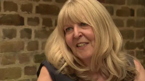Jane smiles off camera. She has blonde wavy hair and is wearing a sleeveless black dress. She sits in front of a brick wall. 