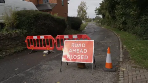 Shaun Whitmore/BBC A red road closure sign and a pair of barriers block access to a closed rural road.