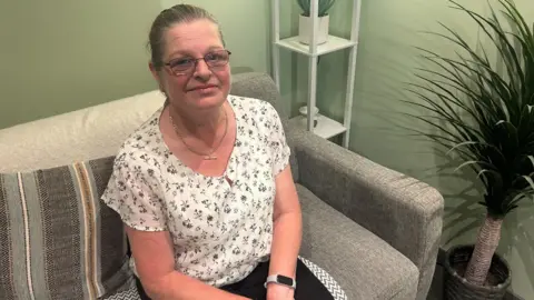 A woman sits on a sofa looking up into the camera. There's a small potted tree beside her. She's wearing a white floral top and a smart watch.