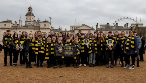 Scotty’s Little Soldiers A large group of young people, and several adults, standing outside a building in London, with the London Eye in the distance. They are all wearing black and yellow scarves. Three people are holding up a banner and two are holding paddles. They are all smiling and looking straight at the camera. 