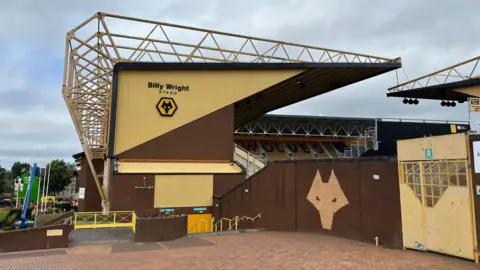 Molineux photographed from outside the stadium, with the Billy Wright Stand, with those words on the side, being most prominent. Part of another stand, which includes the word Wolves by using black seats to spell it out, is in the background.