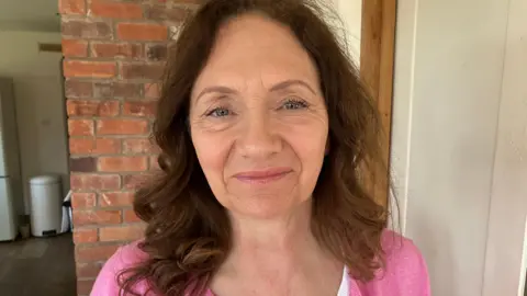 A woman with long shoulder length brown hair smiles at the camera while wearing a pink cardigan and stood in a kitchen at her home.