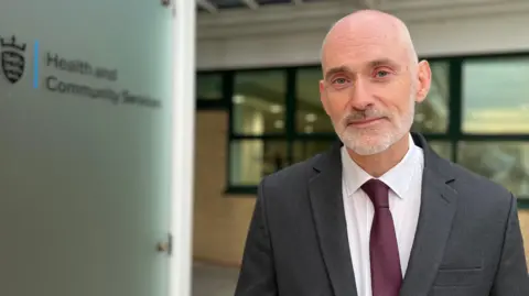 A man stands beside the Health and Community Services building in Jersey. He is wearing a grey suit jacket, white shirt and burgundy tie.