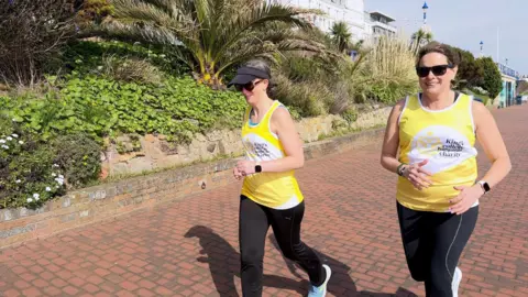 Two women in yellow tops with 'King's College Hospital Charity' on them run along the Eastbourne seafront.