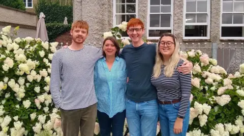 Family Photo Jacqui and her family, stood in front of a flowery bush. They are looking towards the camera and smiling.