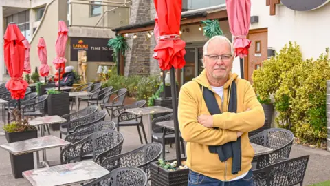 Love Lyme Regis Largigi owner Larry Gibbons stands outside his cafe among the tables and chairs. He is wearing a yellow zipped-up hoody, his arms are folded and he has a serious expression.