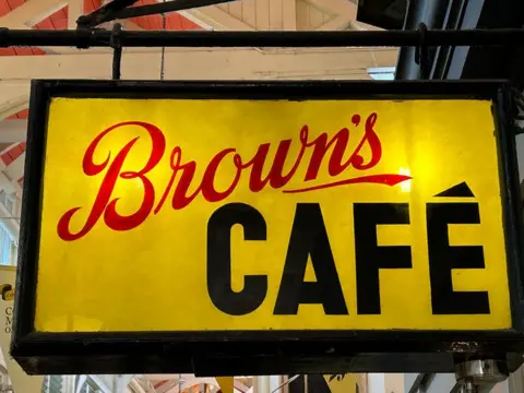 Brown's café sign. The background is yellow with a black outline. The word Brown's is written in red, cursive-style, and the word café is written in black in blocky characters.