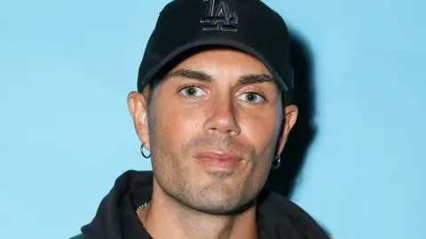 Getty Images Max George wearing a black baseball cap standing against a light blue background in September 2023