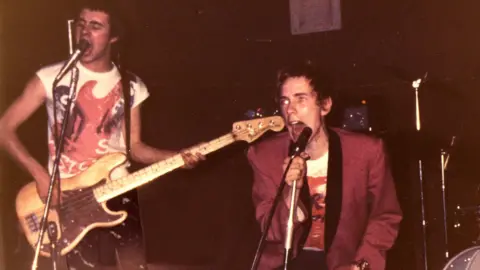 A vintage photo shows two musicians performing energetically on stage with microphones and instruments. One person plays a bass guitar while singing, and the other grips a microphone, leaning forward mid-performance. The dim lighting and grainy texture give the image a raw, punk-rock concert atmosphere.