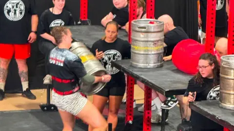 Jade Skill A woman competing in a strongman competition. She is carrying a silver keg and attempting to lift it on to a high shelf. Stewards watch her with one also holding a stop watch.