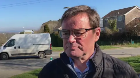 David Landers wearing a black coat with a grey fleece underneath it and a checked shirt. He has glasses and short brown hair. Behind him is a white van pulling out onto the A38.