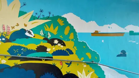 Falmouth BID Part of the mural which features a badger on a cliff and the sea which has two tankers near the horizon and a grey seal with white whiskers closer in. There are large spiky plants and a hedgehog is perched on a stone.