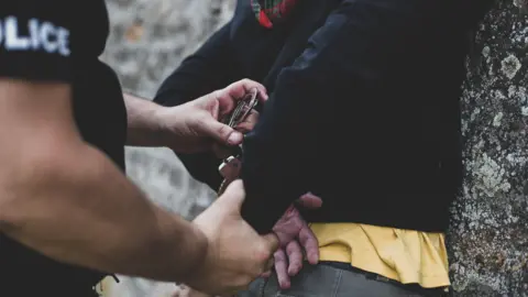 A police officer makes an arrest. The officer's hands and arms are visible as they place handcuffs on to a man who is being pushed up against a wall with his arms behind his back.