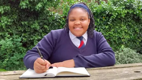 Kinara Dyer-Tuitt has black hair that is sitting on her shoulders. She is wearing a navy headband, a purple V-neck blazer, white shirt and red and blue tie. She is smiling while writing something down in a notebook, sitting on a bench outside.