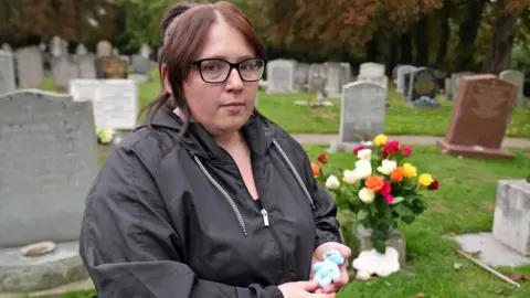 Jasmine Beverley wearing a black coat and black glasses holding her premature baby's small blue teddy bear. She is stood in front of her son's grave which is full of coloured roses.